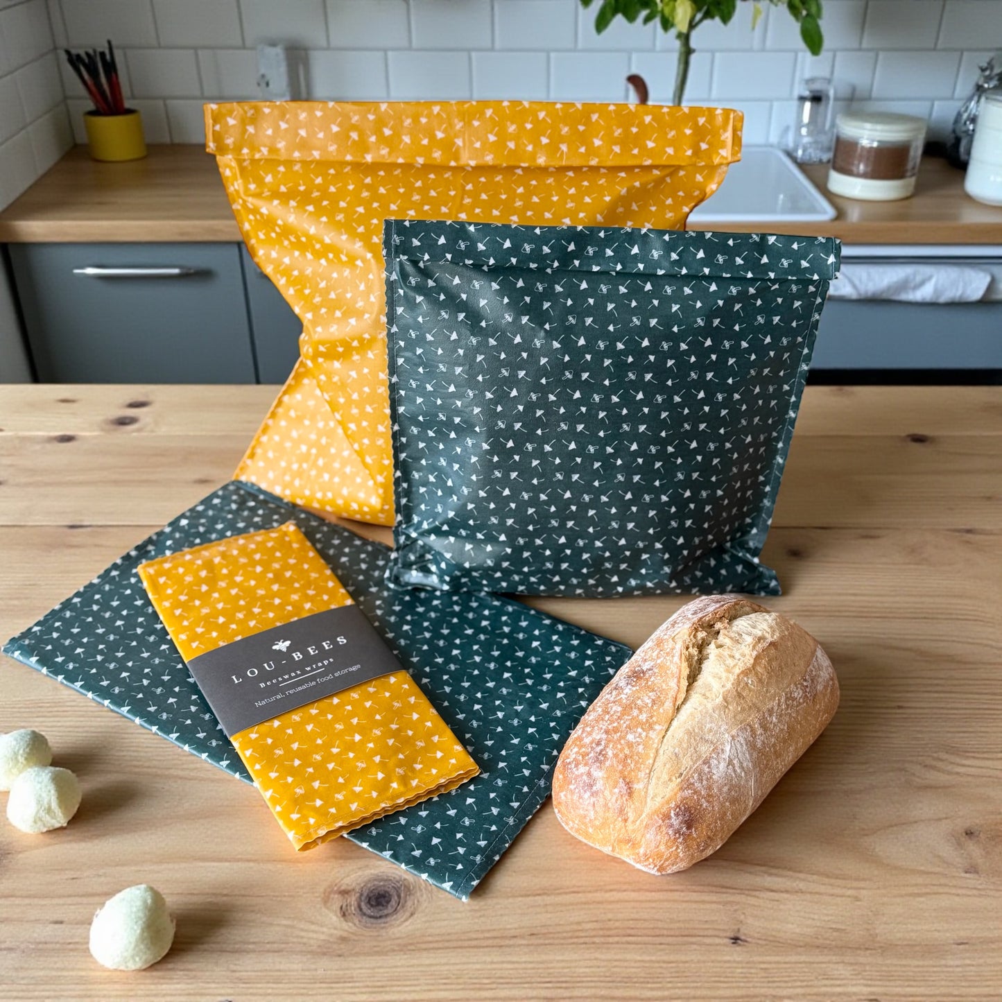 Green and yellow beeswax wraps on a wooden surface with bread and a plant in the background.