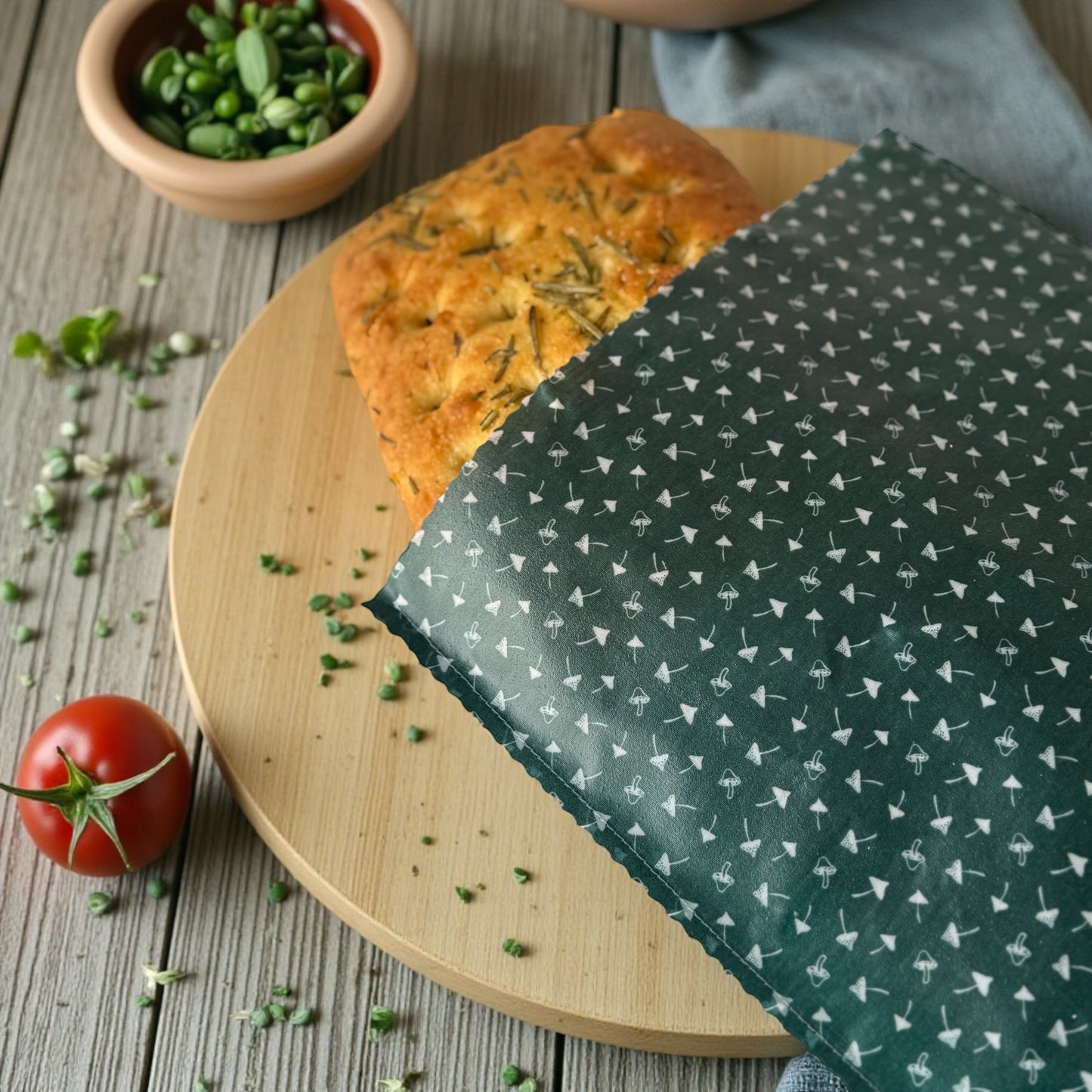 Loaf of bread on a wooden board with a green patterned cloth, surrounded by fresh herbs and a tomato.