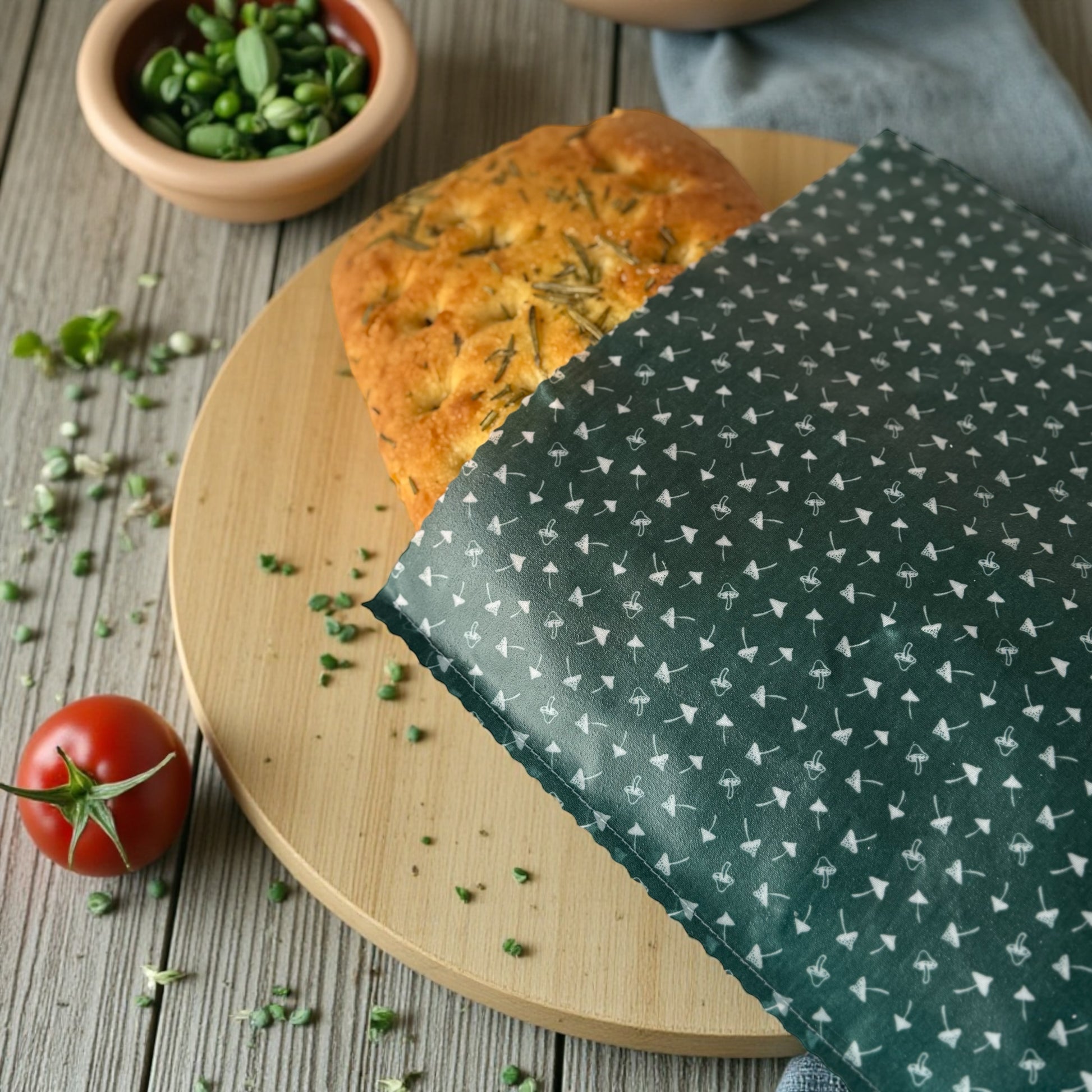 Loaf of bread on a wooden board with a green patterned cloth, surrounded by fresh herbs and a tomato.