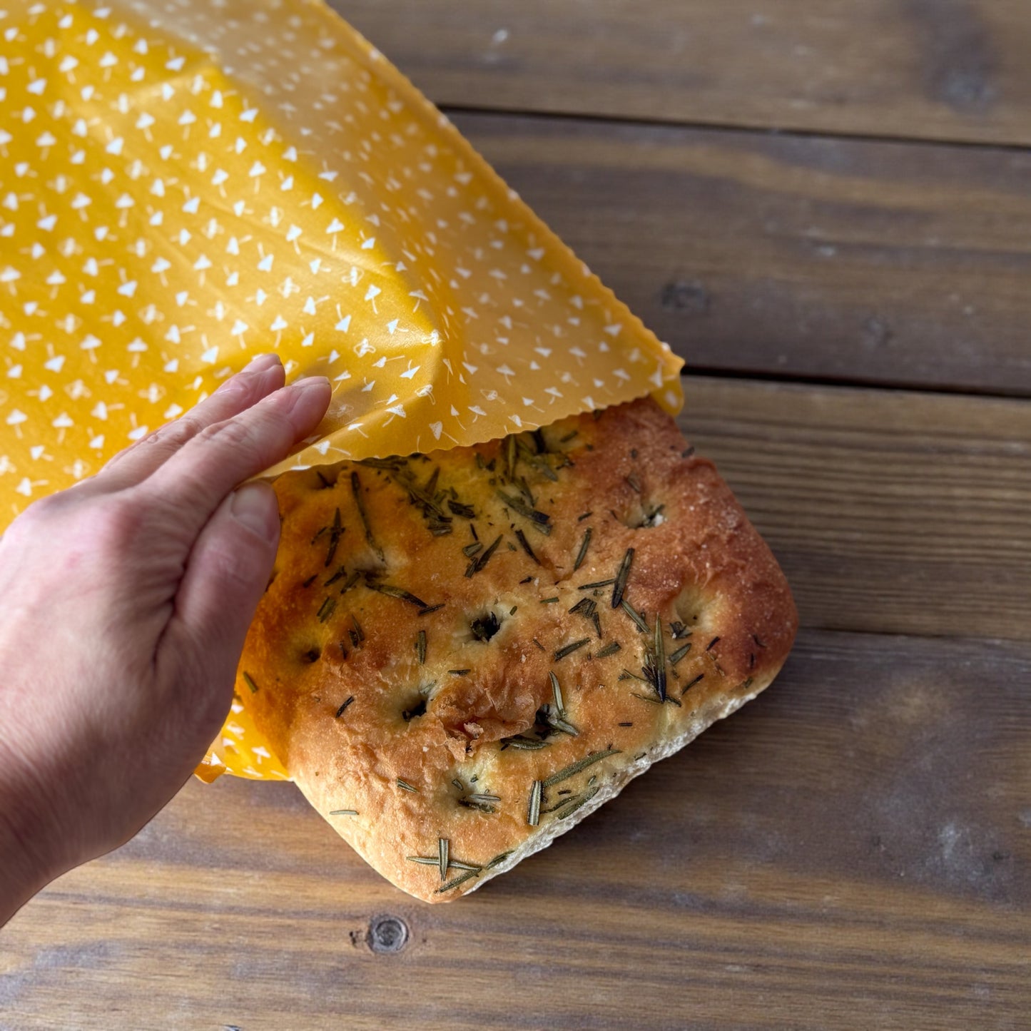 Hand holding a loaf of bread wrapped in yellow beeswax wrap on a wooden surface